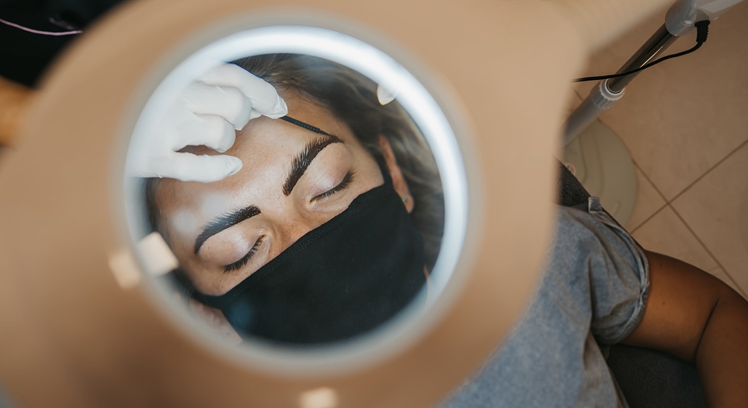 Woman getting eyebrow treatment under magnifying lamp.